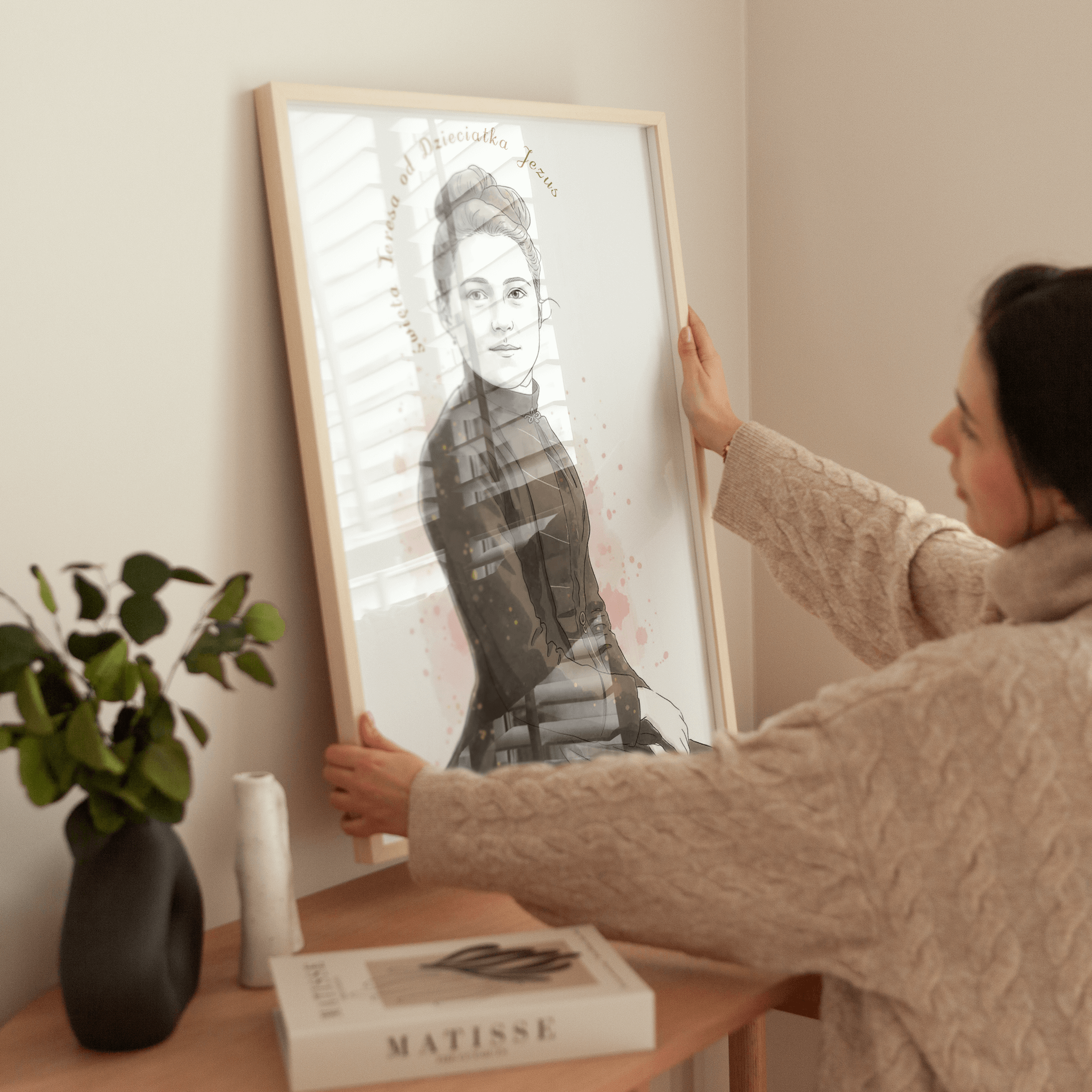 Na zdjęciu widać kobietę wieszającą na ścianie oprawiony portret. Na ilustracji przedstawiona jest młoda kobieta w stylu szkicu artystycznego, a nad jej wizerunkiem znajduje się napis „Święta Teresa od Dzieciątka Jezus”.